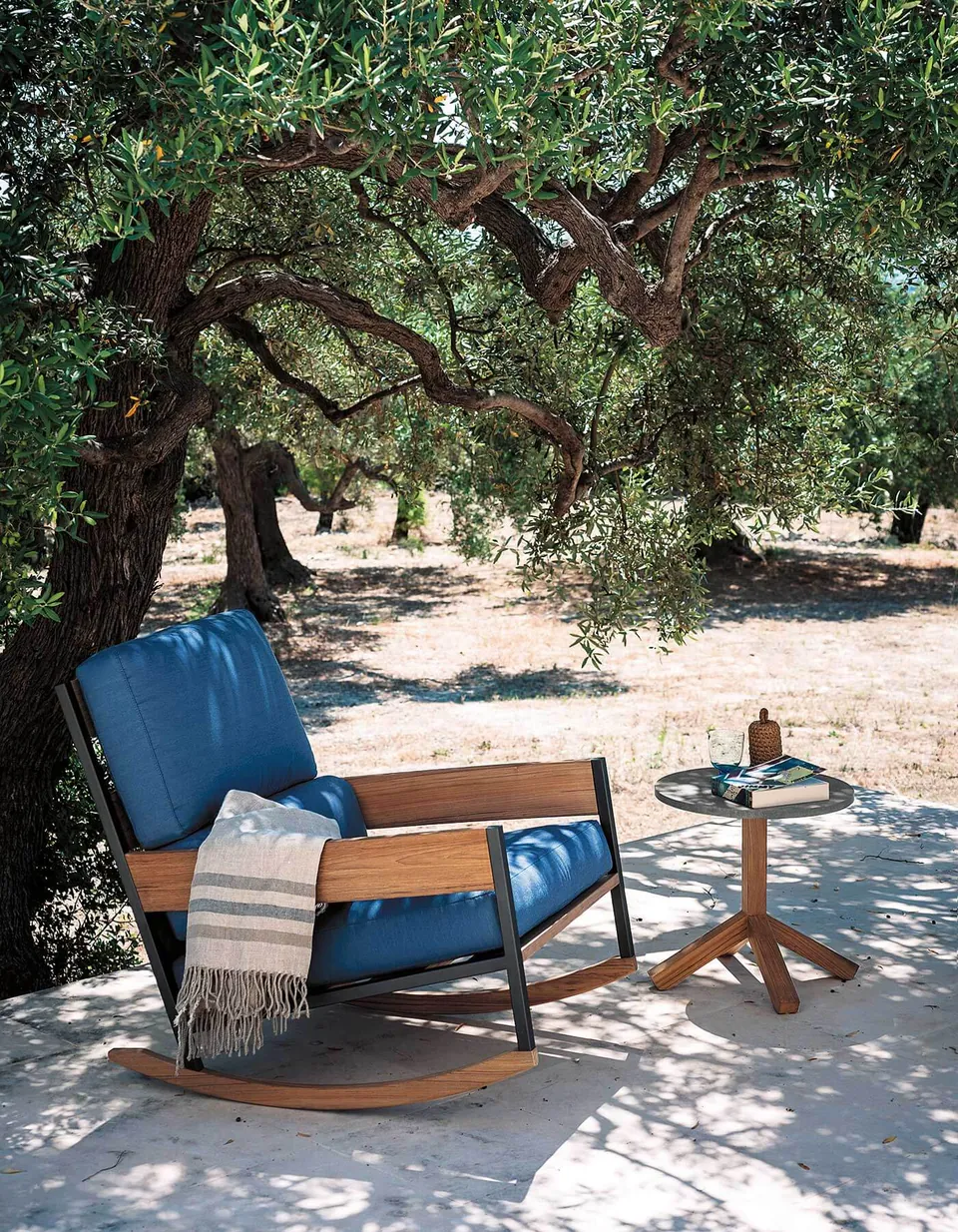 RODA Nap teak rocking chair in a garden under a tree, Italian outdoor furniture catalog photograph